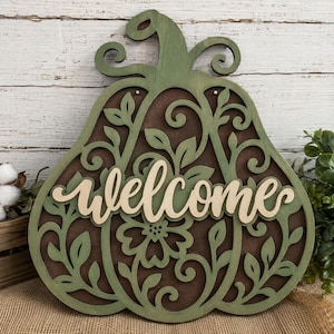 May include: A decorative wooden wall sign shaped like a pumpkin with a sage green laser-cut overlay featuring intricate floral and vine patterns over a dark brown background. The word welcome is written in a light beige script font across the center.
