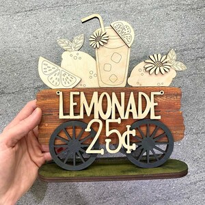 May include: Wooden lemonade stand sign with a glass of lemonade, lemons, and flowers. The sign reads "Lemonade 25¢".