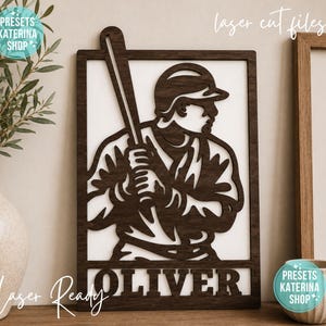 May include: A dark brown laser-cut wooden wall sign featuring a rectangular frame with a baseball player holding a bat in a batting stance. The name OLIVER is cut out in bold capital letters at the bottom of the frame against a white background.