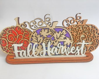May include: Wooden fall harvest sign with three pumpkins in red, purple, and brown with intricate cut-out designs. The pumpkins are layered on top of a wooden base with the words "Fall Harvest" in white.