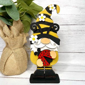 May include: A wooden gnome figurine with a yellow and black striped hat, holding a red ladybug. The gnome has a white beard and is standing on a black base.