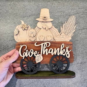 K&ouml;nnte beinhalten: Holz-Thanksgiving-Schild mit einem Truthahn, einem K&uuml;rbis, Mais und einem Kuchen in einem Wagen. Das Schild lautet "Give Thanks".