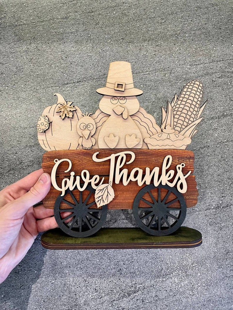 K&ouml;nnte beinhalten: Holz-Thanksgiving-Schild mit einem Truthahn, einem K&uuml;rbis, Mais und einem Kuchen in einem Wagen. Das Schild lautet "Give Thanks".