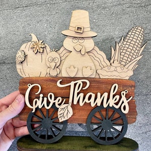 Puede incluir: Señal de Acción de Gracias de madera con un pavo, una calabaza, maíz y un pastel en un carro. El letrero dice "Give Thanks".