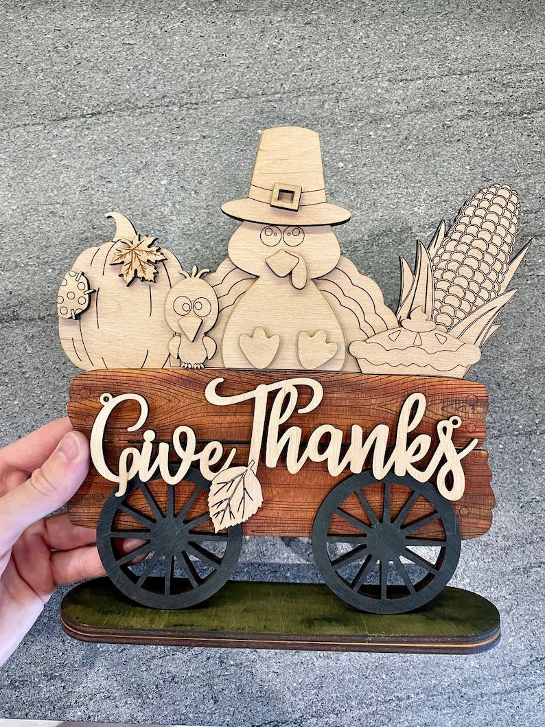 K&ouml;nnte beinhalten: Holz-Thanksgiving-Schild mit einem Truthahn, einem K&uuml;rbis, Mais und einem Kuchen in einem Wagen. Das Schild lautet "Give Thanks".
