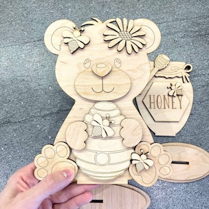 Può includere: Sagoma di legno di un orso che tiene un favo di miele con le api. L'orso ha un fiore sulla testa e un barattolo di miele con la scritta "Honey" sopra.