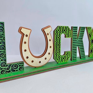Może przedstawiać: Drewniany znak z napisem "LUCKY" w zielonych i brązowych literach z różnymi wzorami. Litera "U" ma kształt podkowy.