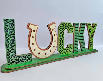 May include: Wooden sign with the word "LUCKY" in green and brown letters with different patterns. The letter "U" is a horseshoe shape.