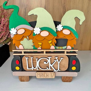 May include: Three gnomes in green hats and brown beards are riding in a black pickup truck. The truck has a wooden sign that says "LUCKY" and "MARCH 17".