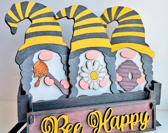 May include: Wooden decorative piece featuring three gnome figures with striped yellow and black hats. Each gnome holds a different bee-themed item. The base has the words "Bee Happy" in yellow script.