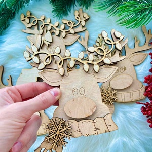May include: Wooden reindeer ornament with a scarf and snowflake design. The reindeer has a light brown color and a string of lights around its antlers.