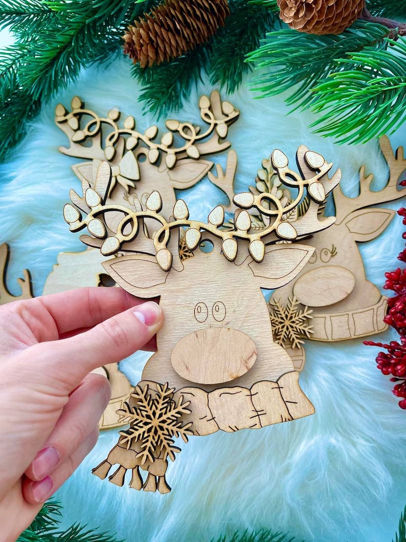 May include: Wooden reindeer ornament with a scarf and snowflake design. The reindeer has a light brown color and a string of lights around its antlers.