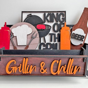 Może przedstawiać: Drewniany znak w stylu grillowania z napisem "Grillin & Chillin" w pomarańczowych literach. Znak jest ozdobiony czarnym grillem, czapką szefa kuchni, butelką piwa i dwiema butelkami ketchupu.