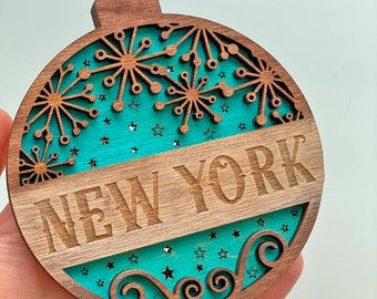 Pode incluir: Um enfeite de Natal de madeira em forma de bola com um fundo azul turquesa e as palavras "NEW YORK" em letras marrons. O enfeite é decorado com um desenho de floco de neve e estrelas.