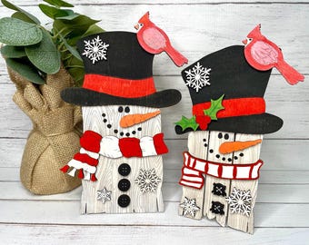 Könnte beinhalten: Zwei Schneemann-Ornamente aus Holz mit schwarzen Zylindern, roten und weißen Schals und Kardinalvögeln obenauf. Die Schneemänner haben Karottennasen, schwarze Knöpfe und weiße Schneeflocken-Akzente.