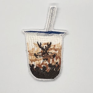 以下が含まれることがあります： 鹿のロゴとストローが付いた茶色と白のボバティーカップの刺繍パッチ。ロゴには「The Alley」と書いてあります