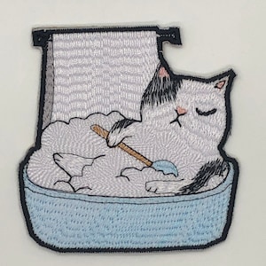 May include: An embroidered patch of a white cat with black markings taking a bath in a blue tub. The cat is holding a blue bar of soap and a wooden stick. The cat has its eyes closed and appears to be relaxing.