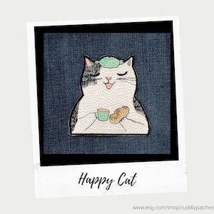 May include: A black and white cat with a green bandage on its head is sitting and holding a green cup and a brown biscuit. The cat is smiling and has its eyes closed. The background is a dark blue denim fabric. The image is titled "Happy Cat" and has the website address "www.etsy.com/shop/cuddlypatches" at the bottom.