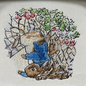 May include: Embroidered image of a brown rabbit wearing a blue jacket and a white hat, hiding behind a bush with red berries.