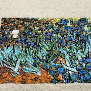 May include: Embroidered artwork depicting a field of blue irises with green leaves and a white flower in the background. The artwork is done in a style reminiscent of Vincent van Gogh.