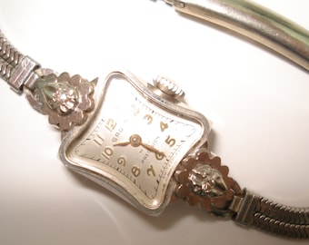 Könnte beinhalten: Eine silberne Gruen Precision Armbanduhr mit weißem Zifferblatt und schwarzen römischen Ziffern. Die Uhr hat ein silbernes Metallband mit einem dekorativen floralen Design an jedem Ende.