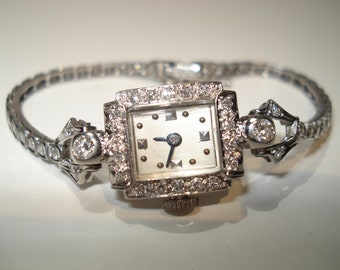 May include: A vintage silver wristwatch with a square face, encrusted with small diamonds. The watch band is also silver and set with small, clear stones. The watch face is white with black hands and markers. The watch has a classic, elegant design.