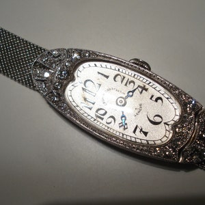 May include: A silver wristwatch with a white face and diamond accents. The watch has a mesh band and the face reads "Girard-Perregaux, Genève".