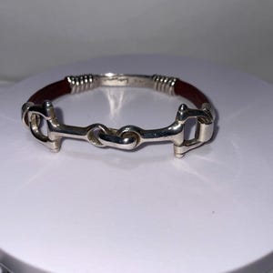 May include: Silver-toned bracelet featuring a horse bit design with a dark red leather band. The bracelet has a polished finish and a classic equestrian theme. The metal components are detailed with intricate designs.