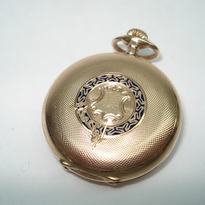 May include: A gold pocket watch with a blue and gold design on the face. The watch has a hinged lid and a chain attachment.