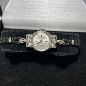 May include: A silver wristwatch with a white face and black hands. The watch is on a black leather strap with silver accents.