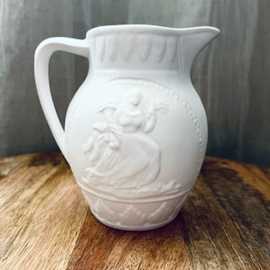May include: A white ceramic pitcher with a handle and spout. The pitcher features a raised relief design of a classical figure in a circular frame. The pitcher is sitting on a wooden surface.
