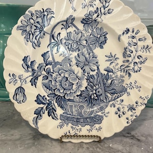 May include: A cream-colored ceramic plate with a scalloped edge featuring a detailed blue transferware pattern of a floral arrangement in a woven basket. The design includes various blooming flowers and foliage extending across the surface.