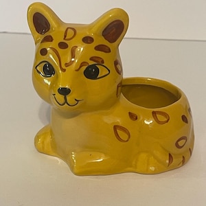 May include: A yellow ceramic planter shaped like a sitting leopard with brown painted spots, dark brown ears, and black eyes. The leopard has a smiling expression and a hollow opening on its back for holding small items or a plant.