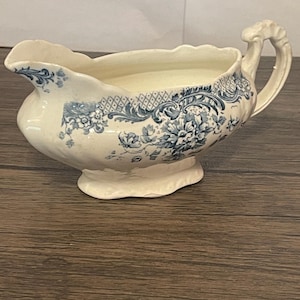 May include: A cream-colored ceramic gravy boat featuring a blue floral transferware pattern. The vessel has a scalloped rim, a curved handle with molded detailing, and a wide pouring spout. The exterior is decorated with intricate blue floral motifs.