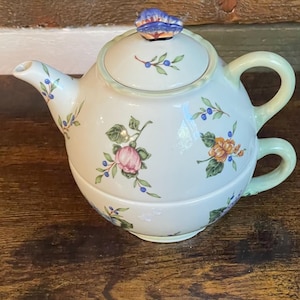May include: A ceramic tea-for-one set featuring a teapot stacked on a teacup. The white surface is decorated with painted pink and orange flowers, green leaves, and small blue berries. A blue butterfly-shaped knob sits atop the teapot lid.