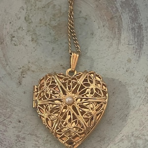Op de afbeelding: Een goudkleurige hartvormige medaillonhanger aan een bijpassende goudkleurige ketting. Het medaillon heeft een ingewikkeld, opengewerkt filigraanontwerp met een kleine, centraal geplaatste witte parel. Het metalen oppervlak is gepolijst.