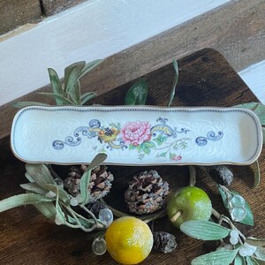 May include: A long, rectangular white ceramic tray with rounded corners and a gold-toned rim. The center features a colorful floral design with a pink peony, a cornucopia filled with fruit, and blue scrollwork patterns on a white background.