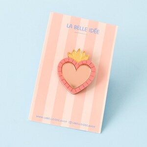 Peut inclure: Épingle en forme de cœur rose avec un motif de couronne dorée, posée sur une carte à rayures roses. La carte porte le texte "LA BELLE IDÉE" et des informations sur le site web. Accessoire décoratif.
