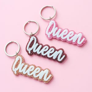 Op de afbeelding: Drie sleutelhangers met zilveren sleutelringen en glitterbehangen bedels die "Queen" in witte letters zeggen. De bedels zijn roze, roségoud en bordeauxrood.