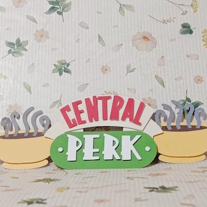 May include: A decorative sign featuring the "Central Perk" logo from the TV show Friends. The sign includes two yellow coffee cups with steam, the words "Central Perk" in red and white, and a green oval with the words "PERK" in white.