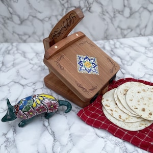 May include: A wooden tortilla press with a carved design and a colorful tile inlay. The press is open and there are several white tortillas on a red and white checkered cloth.
