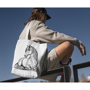 K&ouml;nnte beinhalten: Eine wei&szlig;e Einkaufstasche mit einer schwarzen Linienzeichnung eines Pferdes. Das Pferd liegt mit dem Kopf zur Seite gedreht. Die Tasche wird von einer Person getragen, die ein graues Sweatshirt und Shorts tr&auml;gt.