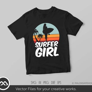 May include: A black t-shirt with a retro-style graphic of a surfer girl standing on a surfboard with palm trees and a sunset in the background. The text "SURFER GIRL" is printed in white below the graphic.