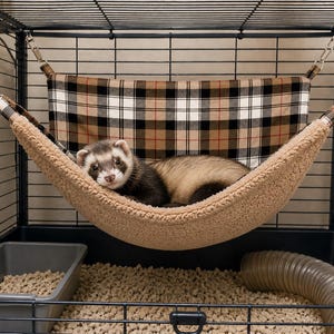 May include: A ferret resting in a hanging hammock suspended by metal clips inside a wire cage. The hammock features a tan, black, and white plaid fabric on the back and a textured, beige fleece lining on the front where the ferret is lying.