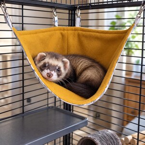 May include: A brown and white ferret resting inside a mustard yellow fabric hammock suspended by metal clips from a black wire cage. The hammock features a soft fleece interior and is attached to the cage frame at four corners with straps.