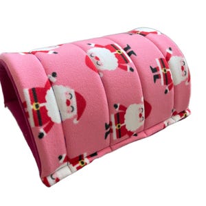 Puede incluir: Cama rosa para mascotas con un estampado de Papá Noel. La cama tiene un diseño curvo y acolchado con una textura suave y afelpada. El estampado de Papá Noel presenta barba blanca, traje rojo y botas negras.