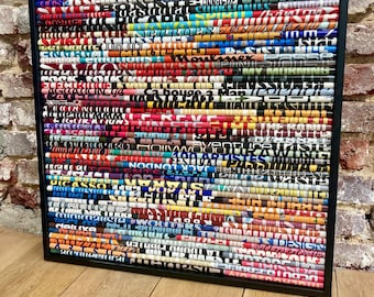 May include: A framed piece of art made from rolled magazine pages, forming a vibrant, textured abstract pattern. The black frame surrounds a collage of colourful text and images from the magazines.