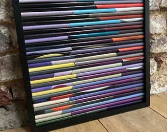 May include: A framed abstract artwork made of colourful, rolled paper strips. The artwork features a geometric pattern of circles and lines in shades of purple, blue, yellow, orange, and grey.