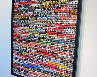 May include: A framed artwork made of rolled up magazine pages, creating a colourful and textured mosaic. The artwork features a variety of words and phrases in black and white, with accents of red, yellow, blue, and green. The frame is black and the artwork is titled "Cafe Racer".