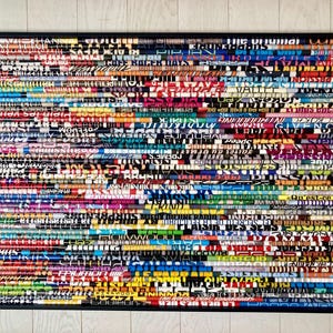 May include: A framed artwork made of colourful magazine strips, each with different text printed on it. The strips are arranged horizontally, creating a layered effect. The frame is black and the background is white.